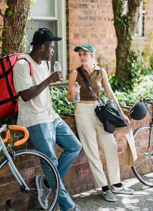 Two people stand beside a bicycle and a brick wall, engaged in conversation while holding water bottles.