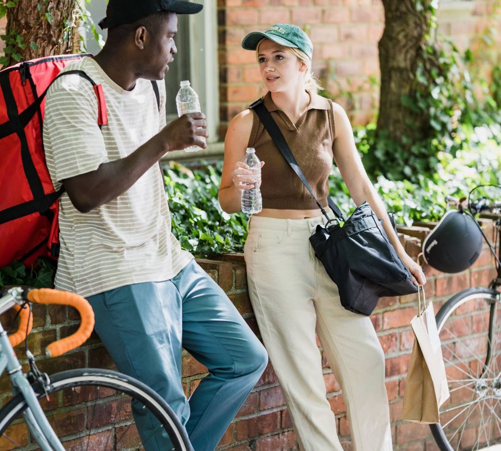 Two people stand beside a bicycle and a brick wall, engaged in conversation while holding water bottles.