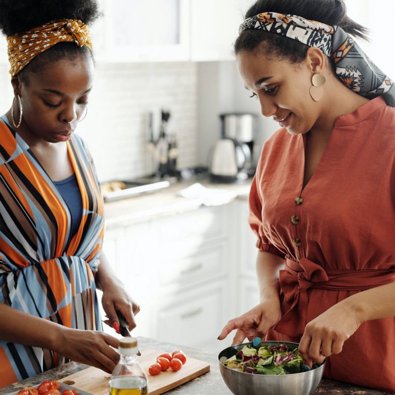 Two women are preparing a salad together in a bright kitchen, one is cutting tomatoes while the other mixes the salad.